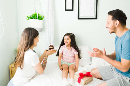 Happy birthday to you. Beautiful hispanic parents singing happy birthday in the morning to their adorable little daughter in the bedroomの写真素材