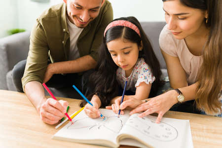 Loving parents teaching her little daughter how to paint a coloring book. Elementary girl learning to color with her mom and dad at homeの写真素材