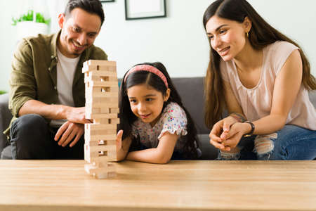 Focused little girl trying to pull a wooden block of a board game tower and trying her move. Latin parents having fun with her daughterの写真素材