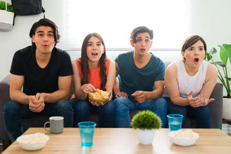 Surprised college friends watching a thriller movie or tv series on the sofa and feeling shocked while eating chipsの写真素材