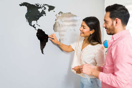 Beautiful attractive couple checking out a world map on the wall of a travel agency office and pinning a country they visit togetherの写真素材