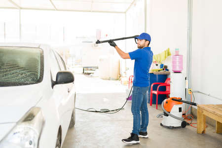 Handsome male worker using a water hose and soap to wash a white vehicle on a car wash. Latin young man cleaning a carの写真素材