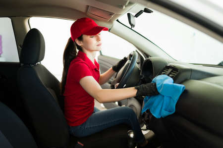 Caucasian female worker sitting in the driver's seat of a customer's car and cleaning with a wash cloth the interior dashboardの写真素材