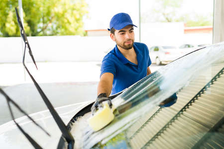 Handsome young man washing a car windshield with soap and a sponge at the car wash and auto detail serviceの写真素材