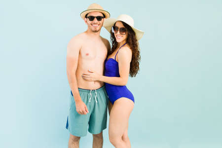 Boyfriend and girlfriend with summer hats and sunglasses smiling and feeling happy on their beach vacations. Attractive couple in swimwear posing and making eye contactの写真素材