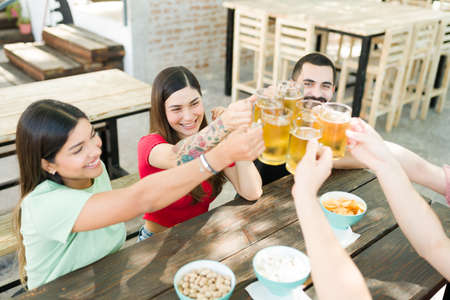 Celebrating our reunion. Excited young friends smiling and making a toast with mugs of beer at a beautiful outdoor barの写真素材