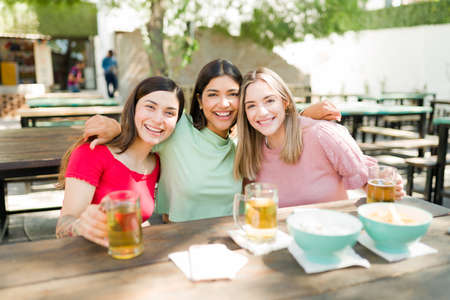 Portrait of excited best friends hugging and smiling while hanging out together and drinking beers. Young women enjoying a fun conversation outdoorsの写真素材