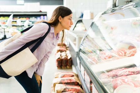 Side view of an attractive woman looking at the freezer with raw meat at the butcher shop or supermarket and deciding what to buy for dinnerの写真素材