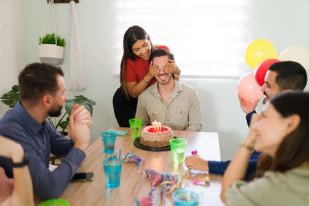 Don't open your eyes. Beautiful young woman covering the eyes of the birthday boy and throwing him a surprise party at homeの写真素材