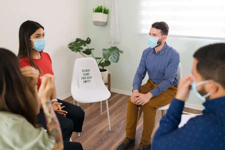 Support group with social distancing. Young people wearing face masks while discussing their...