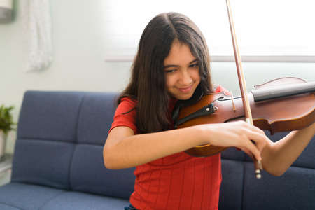 Child playing the violin. Smiling latin girl practicing her music lessons at home. Young student taking online classes to learn to play a new instrumentの写真素材