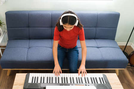 Learning to play the keyboard. High angle of a latin elementary girl with headphones playing the piano and practicing her music lessons at homeの写真素材