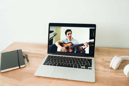 Giving remote music lessons. Handsome latin teacher on a video call working and playing the acoustic guitar for a studentの写真素材