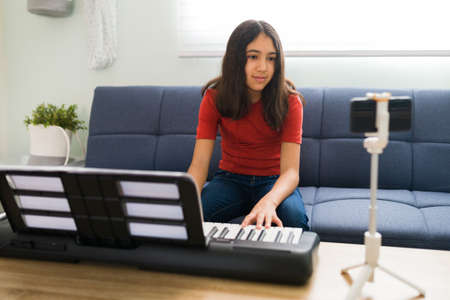 Taking online music classes. Latin preteen girl paying attention to a video call with a piano teacher on her smartphone while sitting on her couch with a keyboardの写真素材