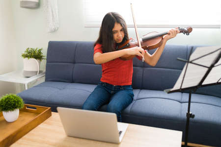 Focused pretty girl following the instructions of her music teacher during an online video call on her laptop and learning to play the violin at homeの写真素材