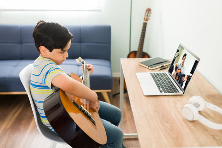 Practicing my acoustic guitar lessons. Caucasian kid sitting on his desk at home and practicing a new song on the guitar during his online music classの写真素材
