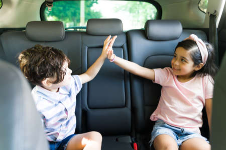 Give me a high five! Beautiful little boy and elementary latin girl having fun and doing a high five while sitting inside the carの写真素材