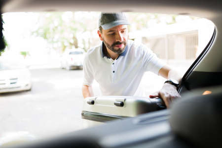 Prepared to relax on a family vacation. Handsome latin man putting luggage in the trunk of the car for their family trip during the holidaysの写真素材