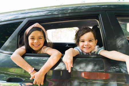 Adorable little brother and sister smiling and making eye contact while putting their heads in the car window. Excited siblings waiting for their parents for a family tripの写真素材