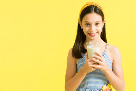 I love milk. Beautiful young girl holding a glass of delicious milk and smiling. Preteen with a hair bow drinking milk against a bright yellow backgroundの写真素材