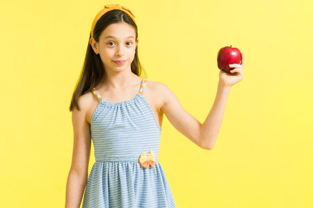 Caucasian little girl eating a red apple as a healthy snack. Happy young girl holding a fruit while making eye contact in front of a bright backgroundの写真素材