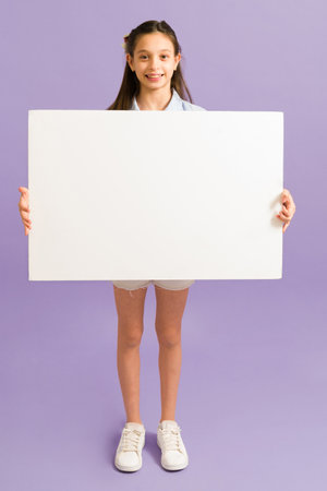 Cute preteen girl wearing a casual outfit with shorts and sneakers while showing a big blank sign to make an announcementの写真素材
