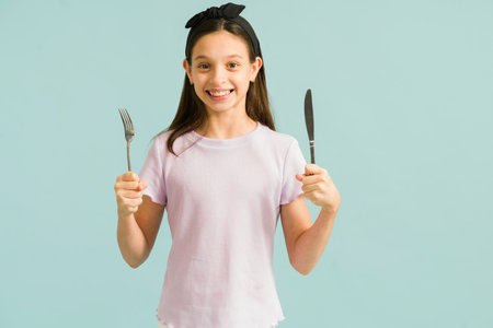 Ready to eat. Hungry little girl smiling while holding a fork and a knife. Beautiful girl preparing...