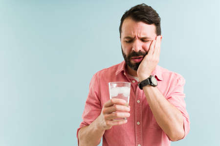 Stressed young man drinking cold water with ice and suffering with tooth sensitivity. Latin man rubbing the side of his faceの写真素材