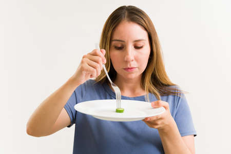 Unhealthy woman in her 20s holding a plate and a fork with very little food because of her bad eating disordersの写真素材
