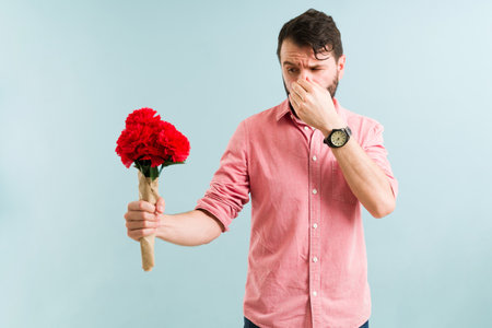 Good-looking man covering his nose while holding a bouquet of red flowers. Latin man having an allergic reaction to pollenの写真素材