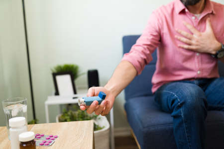 Close up of an adult man having difficulty breathing and holding a preventer inhaler during an asthma attack at homeの写真素材