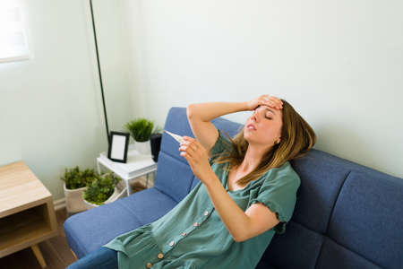 Adult woman with a fever looking at the high temperature on the thermometer at home and thinking about not going to workの写真素材