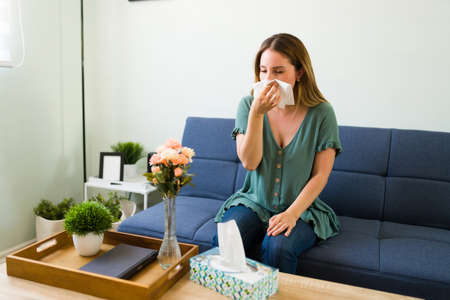 Sick woman with the flu blowing her nose with a tissue. Young woman suffering from a cold and runny nose while feeling unwell at her homeの写真素材