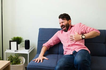 Suffering man with bad chest pain having a heart attack while sitting on the couch. Latin man suffering from coronary issuesの写真素材