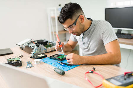 Focused attractive man using a soldering iron to fix broken hardware at the repair shop. Technician...