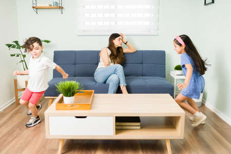 Tired young mom feeling angry and exhausted at home because of her misbehaved children. Little daughter and son playing and running around at homeの写真素材