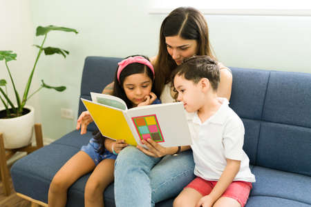 Focused young siblings reading a children's book with her adorable mother before going to bed