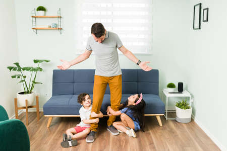 Please dad, don't go. Hispanic dad doesn't know what to do. Small son and daughter holding their...