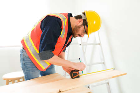Hispanic carpenter in his 30s using a measuring tape and getting ready to cut a wooden panel on the...