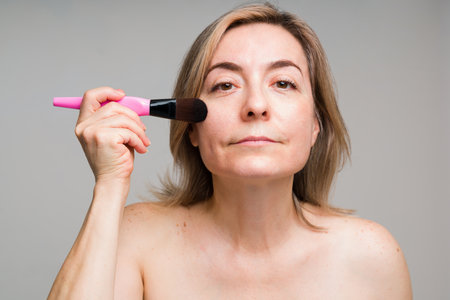 Beautiful mature woman getting ready and putting on foundation with a makeup brush while making eye contactの写真素材