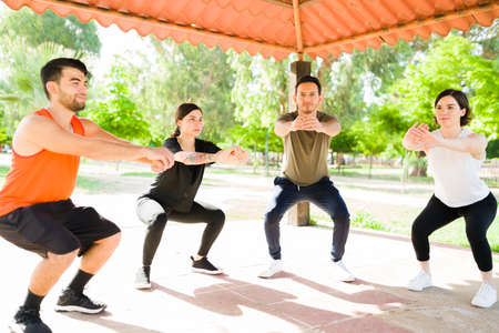 Good-looking young men and women doing squats at the park. Group of friends in activewear doing a...