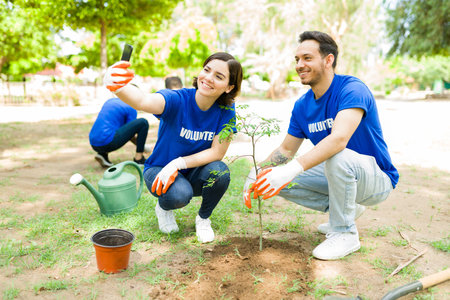 Posting this on social media. Beautiful young woman and man taking a selfie after planting a new tree at the park for community workの写真素材