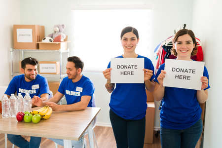 Portrait of two happy women holding signs to donate here. Female volunteers smiling and asking...