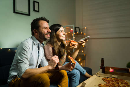 Hispanic husband and wife laughing and watching a comedy movie while eating dinner together for a...