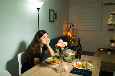 He stood me up. Sad young girlfriend feeling upset while waiting for her partner to show up for a dinner date at homeの写真素材