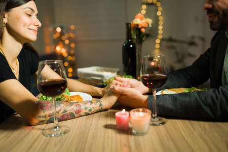 Close up of a couple in love holding hands and having a romantic moment while enjoying a fancy evening date at home for their anniversaryの写真素材