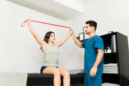 Attractive young woman using a resistance band with her latin male physiotherapist while doing...