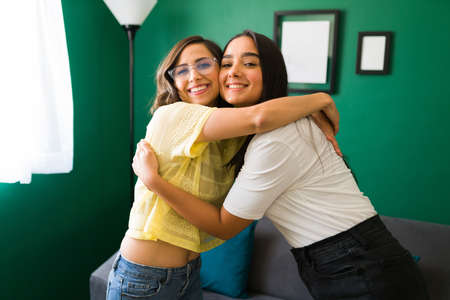Portrait of two beautiful young women smiling and hugging in their apartment while looking at the...