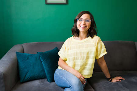Portrait of an hispanic woman with glasses smiling and resting on the couch. Cheerful woman wearing...