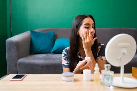 Hispanic gorgeous woman in pajamas sitting in the living room while trying skin care products and a moisturizerの写真素材